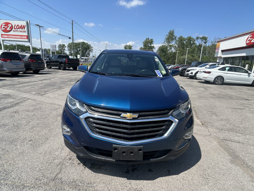 Front view of a blue Chevrolet SUV in a car dealership lot with other vehicles and an EZ LOAN sign in the background, under a blue sky.