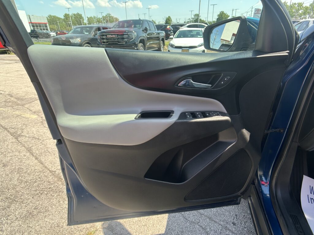 Interior of a car door showing the armrest, window controls, and chrome handle with the side mirror visible; outside is a dealership parking lot with many vehicles.