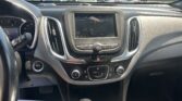 Driver‑side view of a car interior with the center touchscreen on, showing radio info, climate controls, and gear shifter in the foreground, parked cars visible through the windshield.