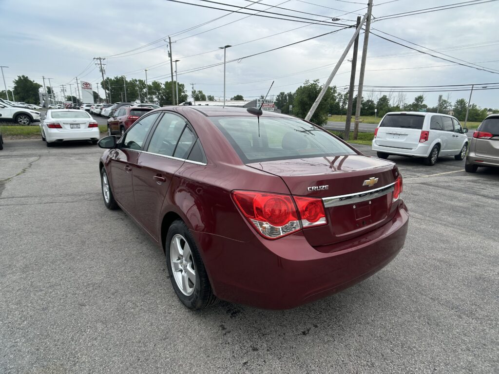 Rear three-quarter view of a maroon Chevrolet Cruze in a parking lot, with white vans and other cars in the background and power lines overhead.
