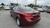 Rear three-quarter view of a maroon Chevrolet Cruze in a parking lot, with white vans and other cars in the background and power lines overhead.