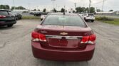 Rear view of a red Chevrolet Cruze LT parked in a car lot with other vehicles and a dealership in the background.