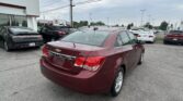 Red Chevrolet sedan with an LT badge in the foreground at a car dealership lot, with rows of cars and a white building on the left.