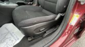 Interior of a red car showing the front passenger/driver area: gear shifter, center console, and black fabric seats with an open door visible at the edge. Includes a floor mat with partial text.