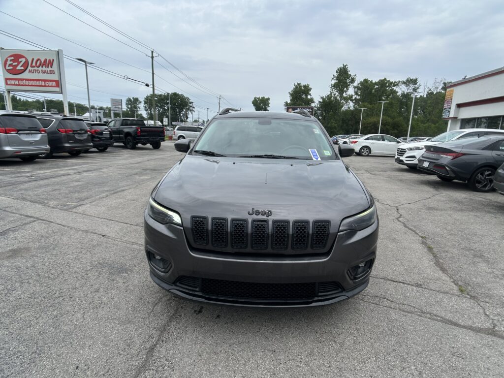 Front view of a gray Jeep SUV in a car lot with a dealership sign on the left and rows of vehicles surrounding it.