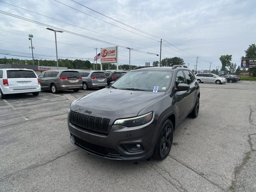 Gray Jeep SUV in the foreground at a car dealership lot with a row of minivans and an EZ LOAN sign in the distance under a cloudy sky.