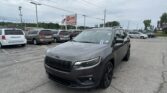Gray Jeep SUV in the foreground at a car dealership lot with a row of minivans and an EZ LOAN sign in the distance under a cloudy sky.