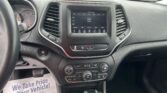 Interior of a car showing the center touchscreen, climate controls, gear shifter, and steering wheel with a parking lot visible through the windshield.