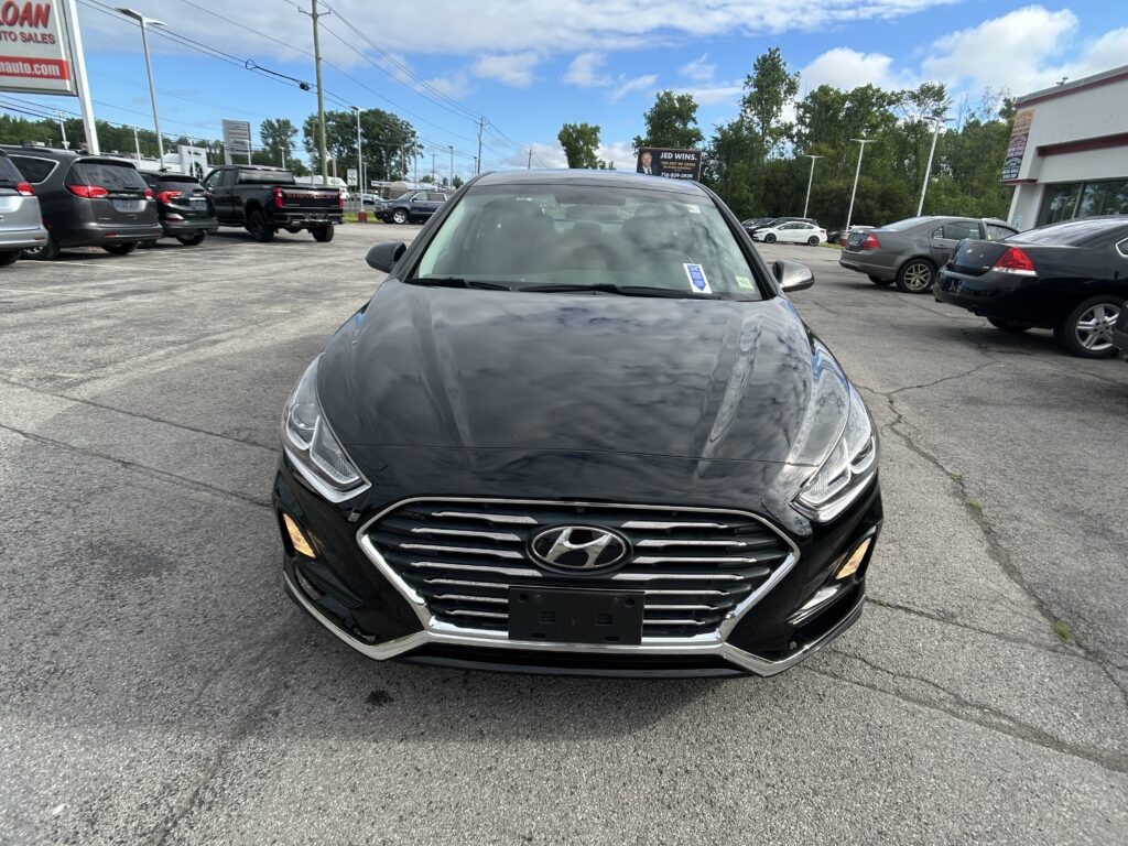 Front view of a black Hyundai SUV in a car dealership lot, with other cars parked nearby and a blue sky above.