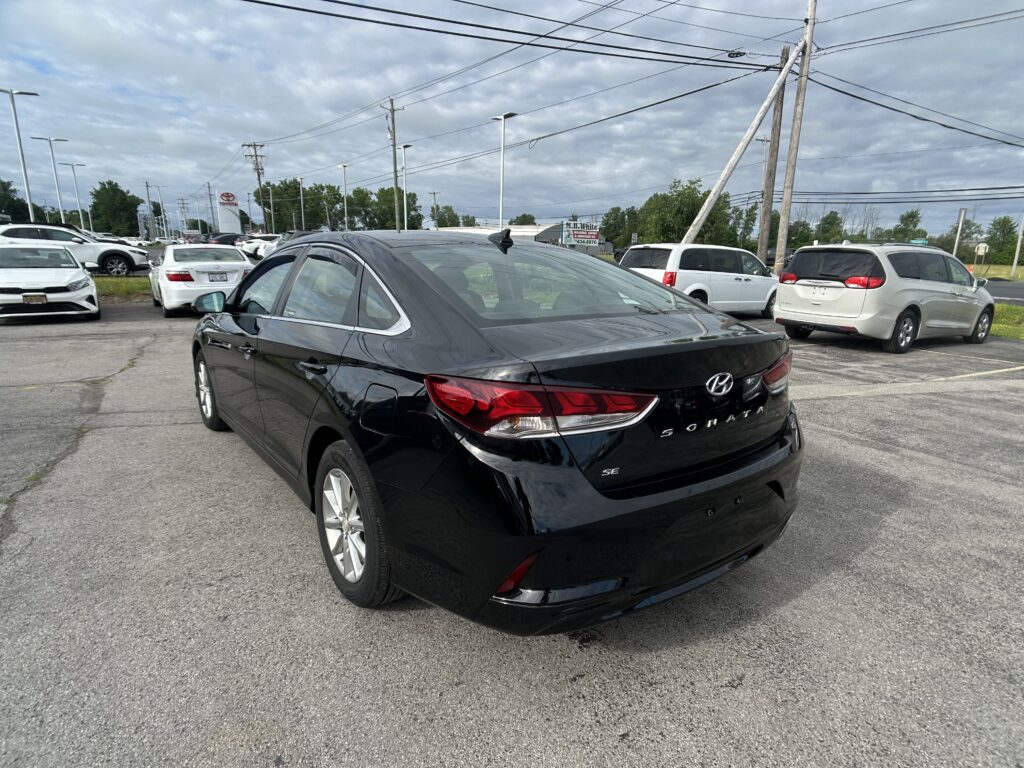Black Hyundai Sonata in the foreground of a crowded car dealership lot with white sedans and SUVs in the background under a cloudy sky.