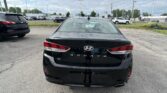 Rear view of a black Hyundai Sonata in a dealership parking lot with other cars and a Toyota sign in the distance.