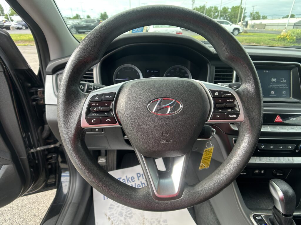 Close-up of a car's steering wheel with dashboard gauges and the center touchscreen visible, seen from the driver’s seat in a parking lot.