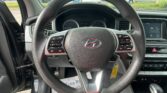 Close-up of a car's steering wheel with dashboard gauges and the center touchscreen visible, seen from the driver’s seat in a parking lot.