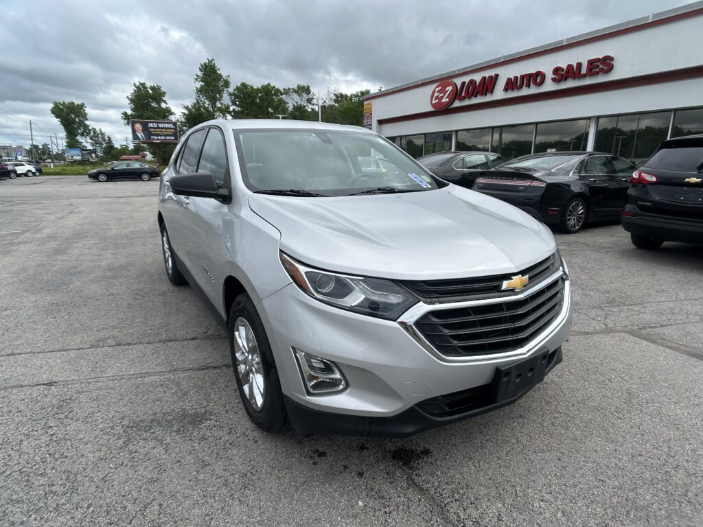 Silver Chevrolet SUV in the foreground at an auto dealership lot, EZ LOAN AUTO SALES visible on the building with other cars parked nearby under a cloudy sky.