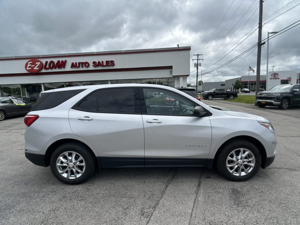 White Chevrolet Equinox parked in a dealership lot with E-Z Loan Auto Sales sign on a building in the background and a Toyota showroom across the street.