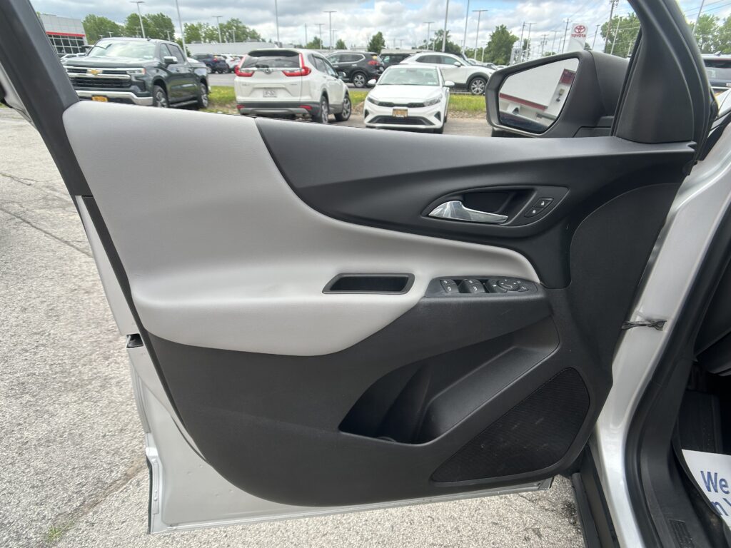 Driver-side car door panel with window controls; view of a busy parking lot outside through the door and mirror.