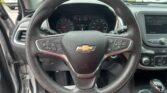 Driver’s-seat view of a car interior: steering wheel with Chevrolet logo, instrument cluster, and right-side infotainment screen; parking lot visible outside.