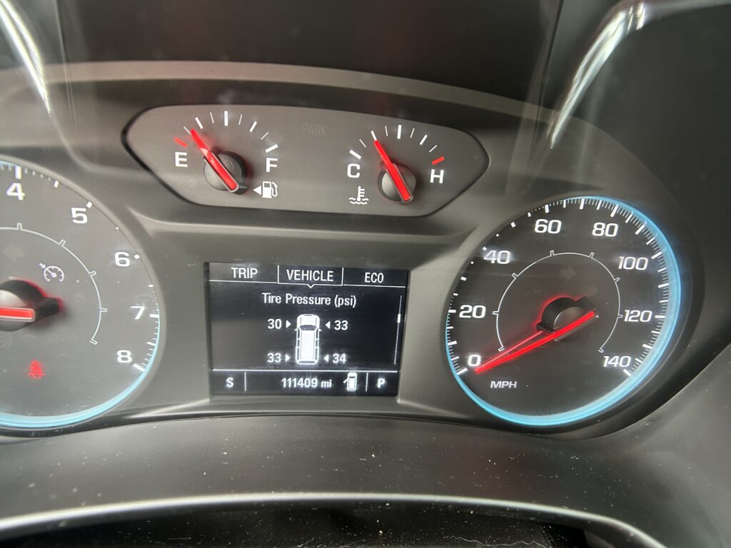 Car dashboard with tachometer on left and speedometer on right, red needles at mid-range, digital display below showing trip info and tire data