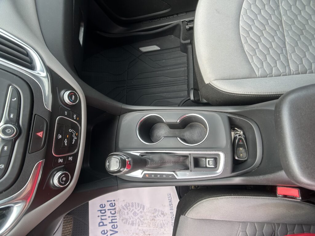Center console of a car interior with gear selector, cup holders, climate controls, and a paper on the floor mat nearby.