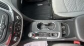 Center console of a car interior with gear selector, cup holders, climate controls, and a paper on the floor mat nearby.