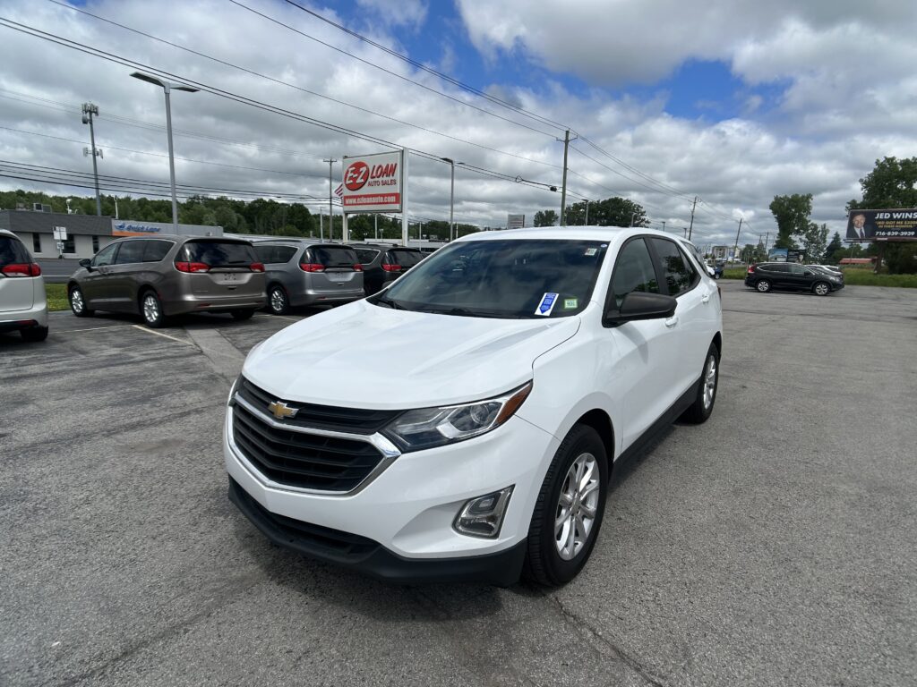 White Chevrolet SUV in foreground at a car lot with rows of minivans, utility poles, and a red EZ LOAN Auto Sales sign in the background under cloudy skies.