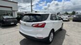 White Chevrolet SUV in a busy car dealership lot, with a white building on the left and a cloudy blue sky above.
