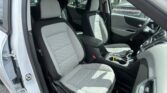 Open car door showing the vehicle's front seats with light gray quilted upholstery and a center console. Steering wheel is on the right side inside. Outside view shows a parking lot.