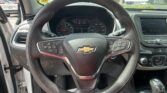 Driver's view of the car interior: steering wheel with Chevrolet emblem, gauge cluster, and center touchscreen; outside parking lot visible through the windshield.