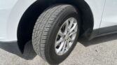 Close-up of a car tire and alloy wheel mounted in the front wheel well of a light-colored vehicle on pavement, showing the tread pattern and rim detail.