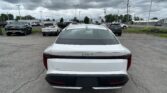 Rear view of a white Kia K5 in a crowded dealership lot under a cloudy sky.