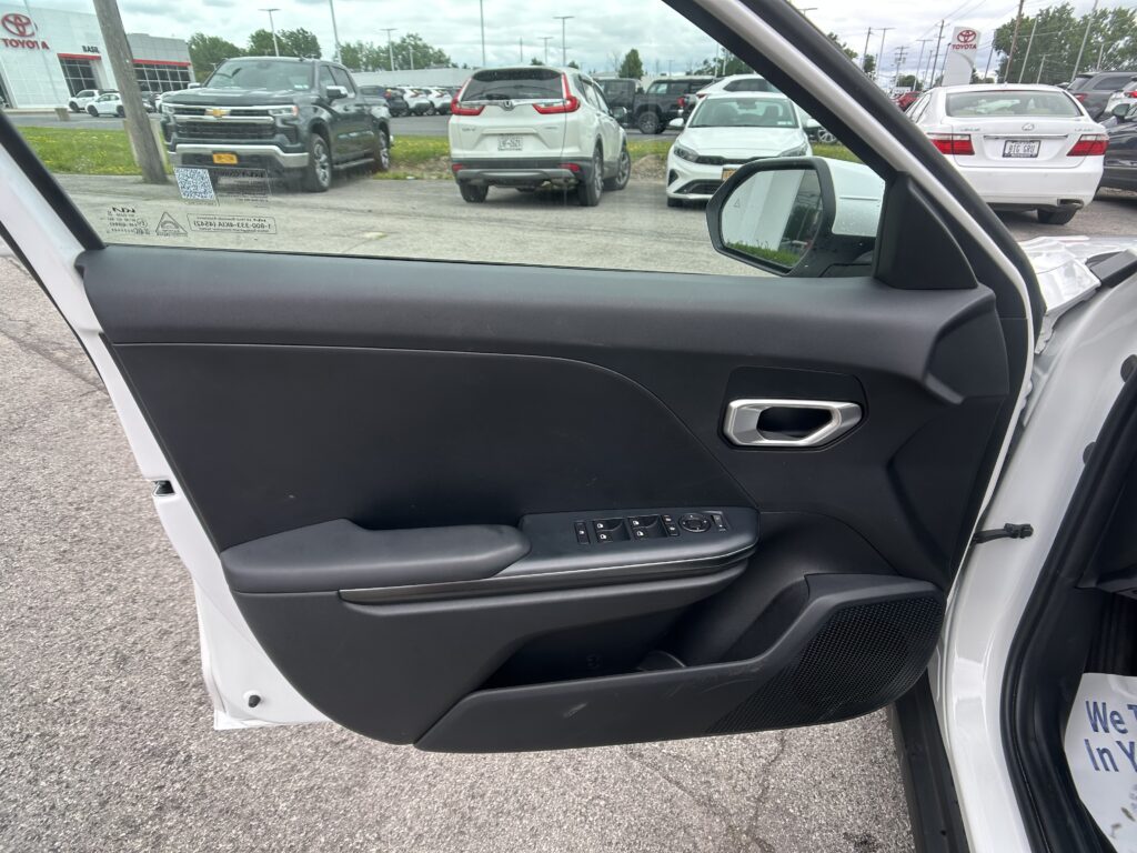Open front passenger door view showing black interior panel and window controls, with a car dealership lot of white and gray SUVs visible outside a white SUV.