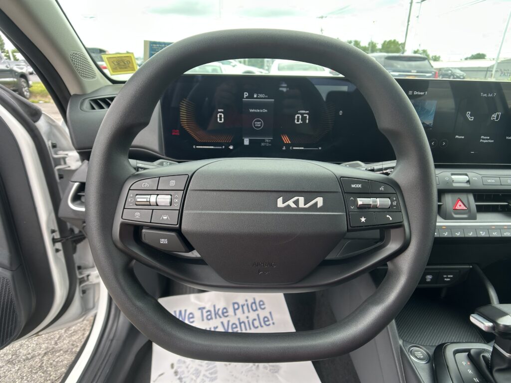 Steering wheel view from the driver’s seat in a Kia, with a digital instrument cluster and parked cars visible outside the windshield.