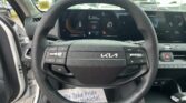 Steering wheel view from the driver’s seat in a Kia, with a digital instrument cluster and parked cars visible outside the windshield.