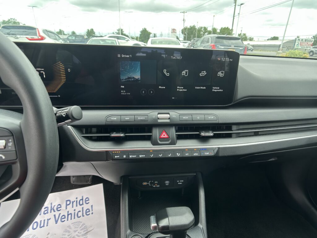 Interior of a car showing a wide horizontal touchscreen displaying media icons, parked in a lot seen through the windshield, with climate controls below.