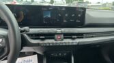 Interior of a car showing a wide horizontal touchscreen displaying media icons, parked in a lot seen through the windshield, with climate controls below.