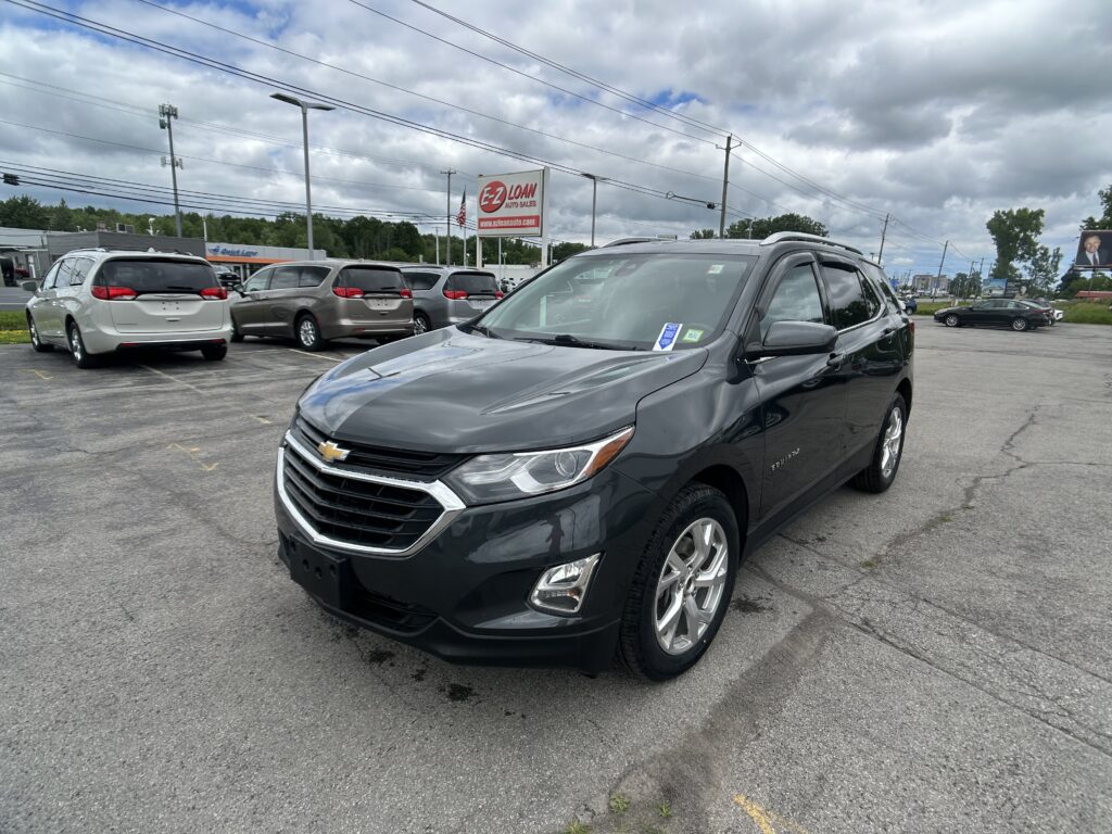 Gray Chevrolet SUV in the foreground parked at a dealership lot with other minivans, a red EZ Loan Auto Sales sign in the background, and a cloudy sky above.