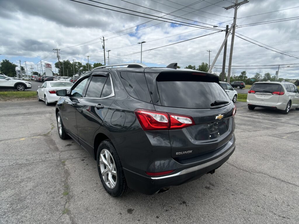 Rear view of a dark gray Chevrolet Equinox SUV in a car dealership lot with other vehicles and power lines in the background under a cloudy sky, at center-right edge of the frame.