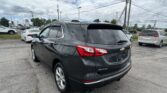 Rear view of a dark gray Chevrolet Equinox SUV in a car dealership lot with other vehicles and power lines in the background under a cloudy sky, at center-right edge of the frame.