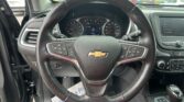Steering wheel and instrument cluster from the driver's seat with a Chevy emblem, dashboard gauges, and a right-side touchscreen; parking lot visible outside.
