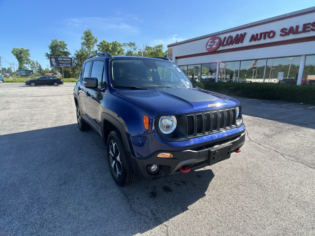 Blue Jeep SUV in the dealership lot with E-Z Loan Auto Sales storefront and large windows in the background, under a bright blue sky.