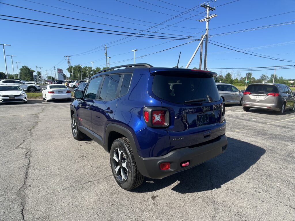 Blue Jeep SUV in a dealership parking lot under a clear blue sky; power lines overhead.