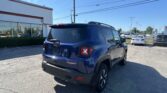 Rear three-quarter view of a blue Jeep SUV parked in a dealership lot with other cars and a white building in the background under a clear blue sky.