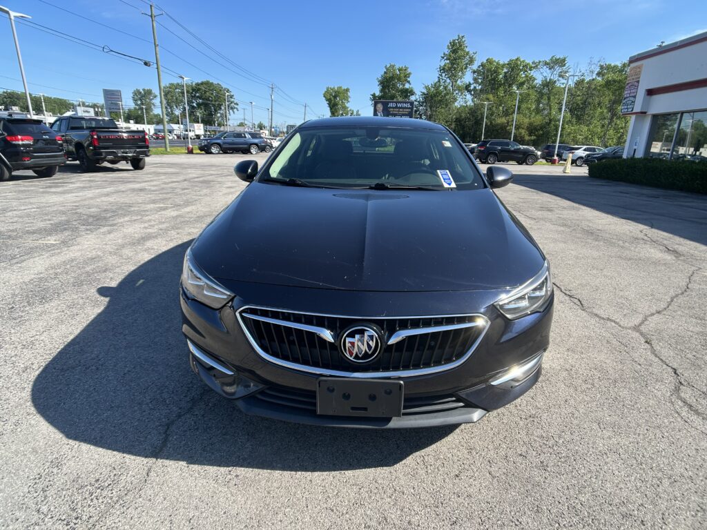 Front view of a dark gray Buick SUV in a dealership lot, with other cars and a building in the background.