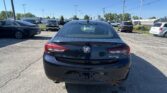 Rear view of a black Buick Regal parked in a car dealership lot with other vehicles and a clear blue sky behind.