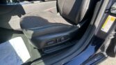 Driver's seat and center console of a dark car interior, showing seat adjustment controls and stitching detail with sunlight on the door frame.