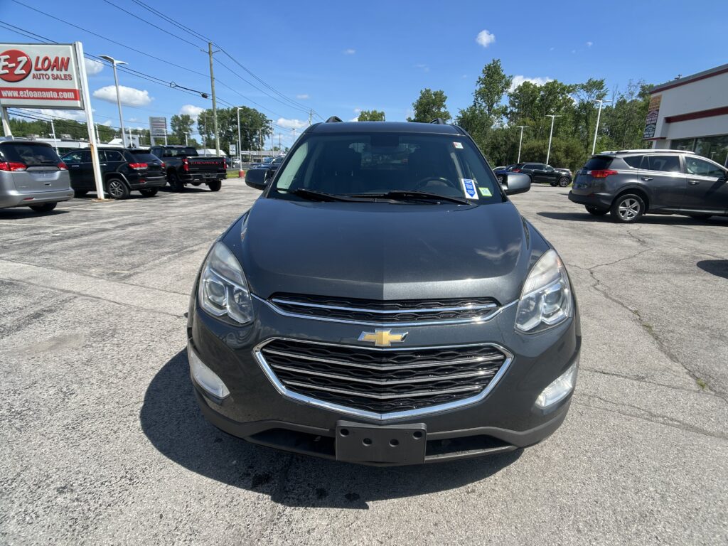 Front view of a gray Chevrolet SUV in a car dealership lot with other vehicles and a large EZ LOAN sign on the left under a clear blue sky.