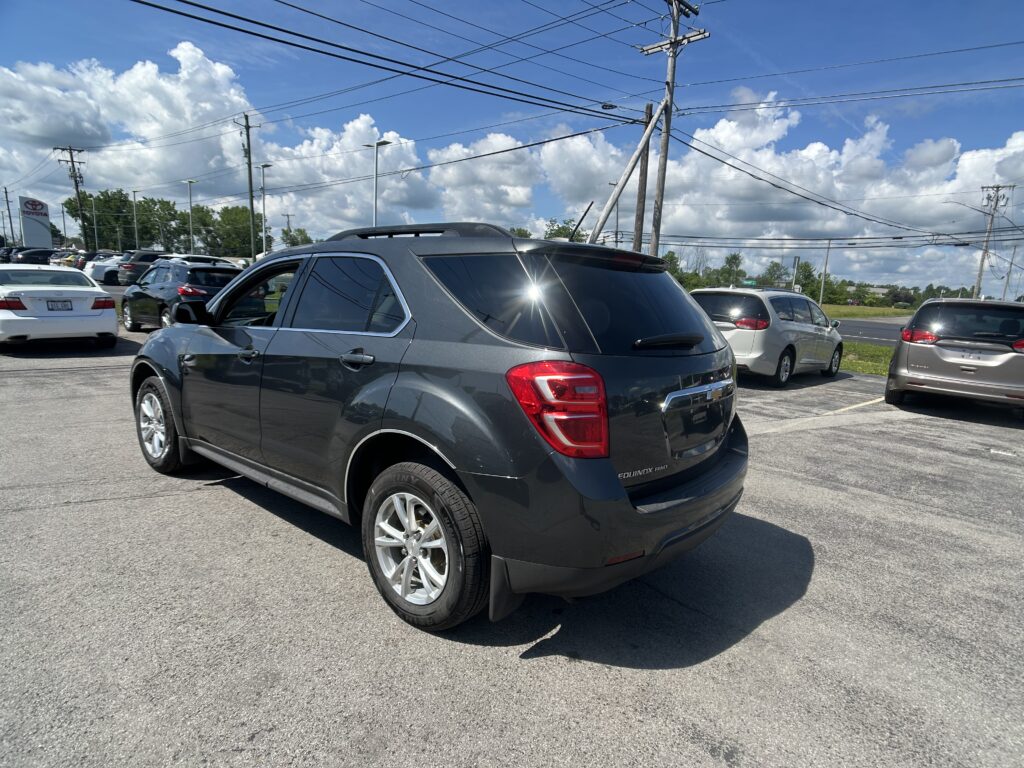 Rear three-quarter view of a gray SUV in a car dealership lot with other cars and power lines overhead against a blue sky with clouds, foreground focus on the vehicle.