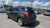 Rear three-quarter view of a gray SUV in a car dealership lot with other cars and power lines overhead against a blue sky with clouds, foreground focus on the vehicle.