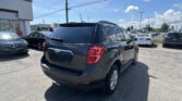 Dark gray Chevrolet SUV in a busy dealership lot on a sunny day, with cars lined up and a white showroom building to the left.