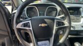 Inside a Chevrolet SUV cockpit with steering wheel center, instrument cluster, and center console; parked cars visible through the windshield outside.
